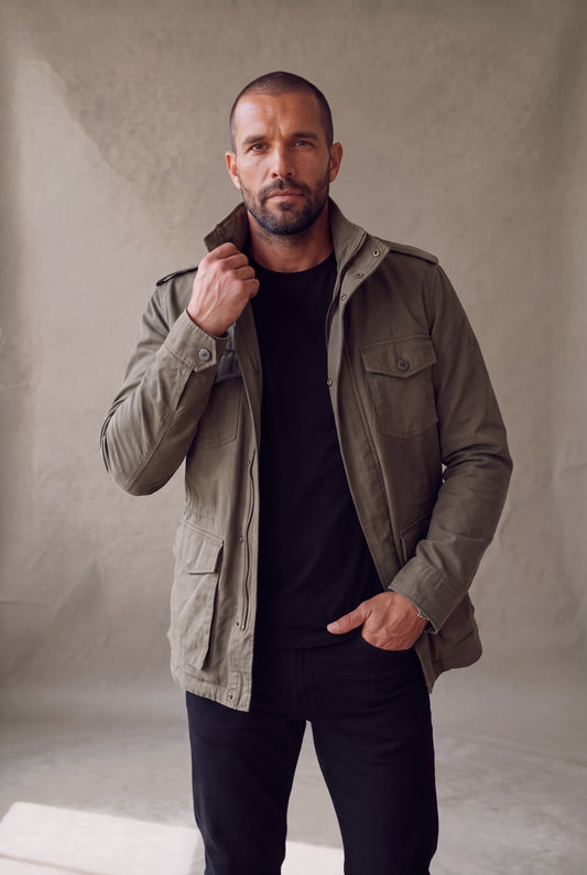 Medio cuerpo de la chaqueta militar verde oliva para hombre Luck and Load: bolsillos de pecho con boton tonal, cuello alto y cierre, sobre camiseta negra.
