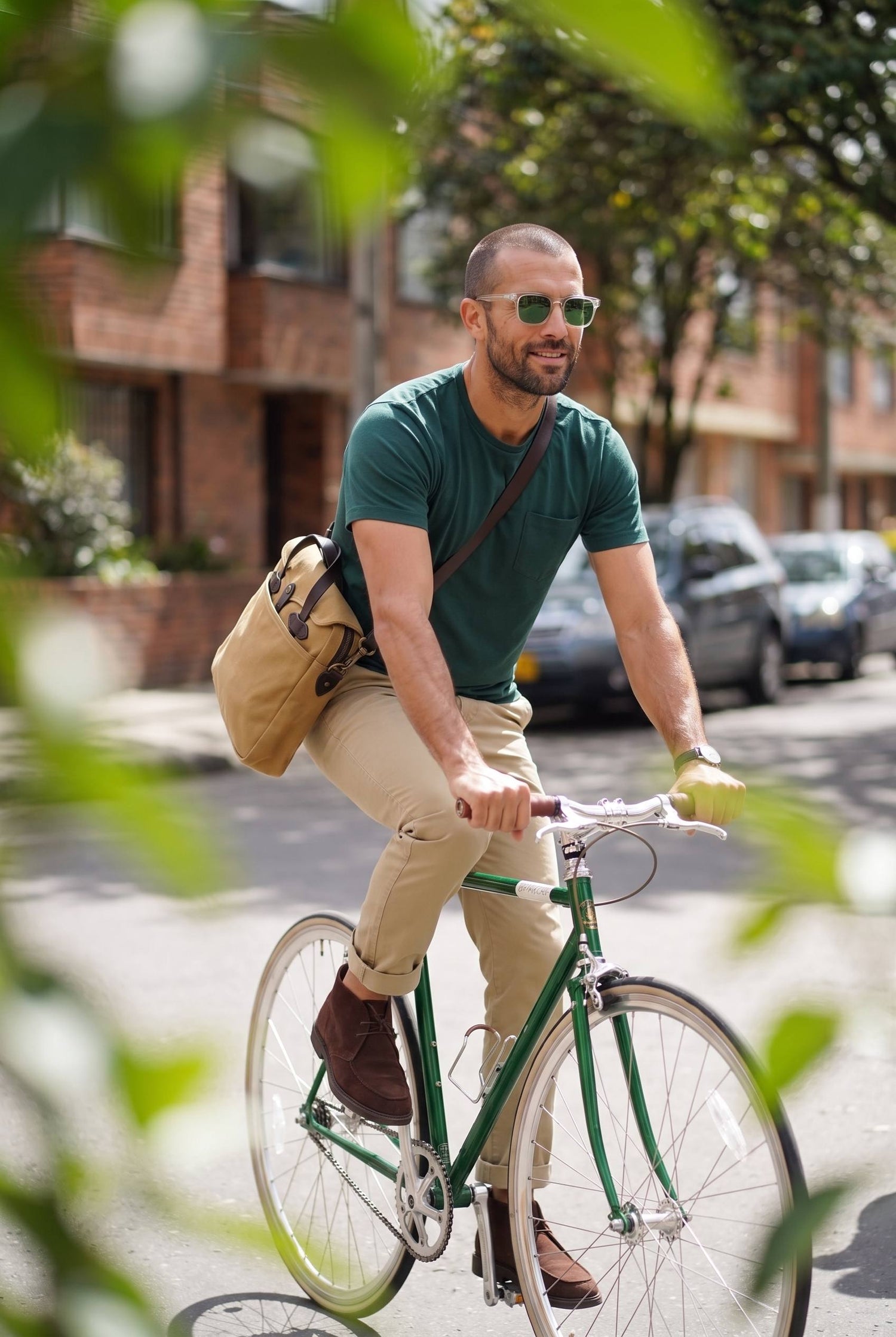Hombre con camiseta verde pino de bolsillo montado en una bicicleta por la ciudad con su maletin, un plan de fin de semana.