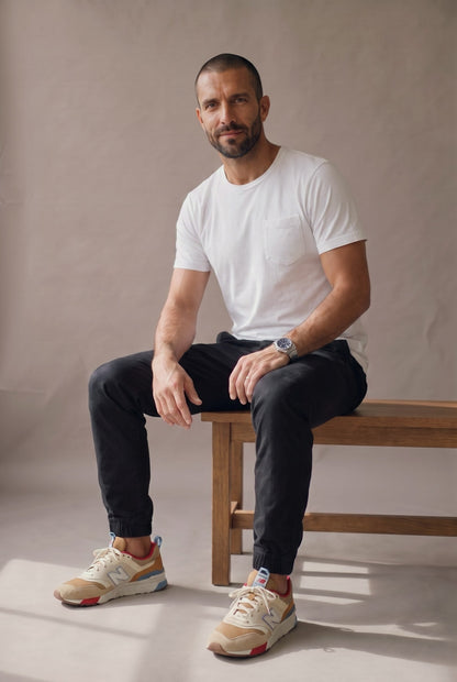 Hombre sentado en una banca con camiseta blanca de bolsillo, jogger negro y tenis, en estudio neutro