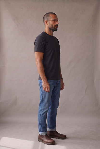 Hombre de perfil con camiseta negra de bolsillo, jean índigo cuffed y botas cafés, en estudio neutro