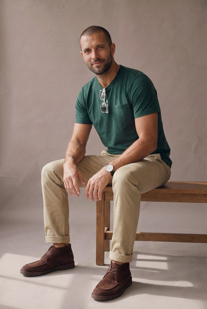 Hombre de perfil derecho con camiseta verde pino de cuello redondo con bolsillo en algodon peruano, en estudio neutro.