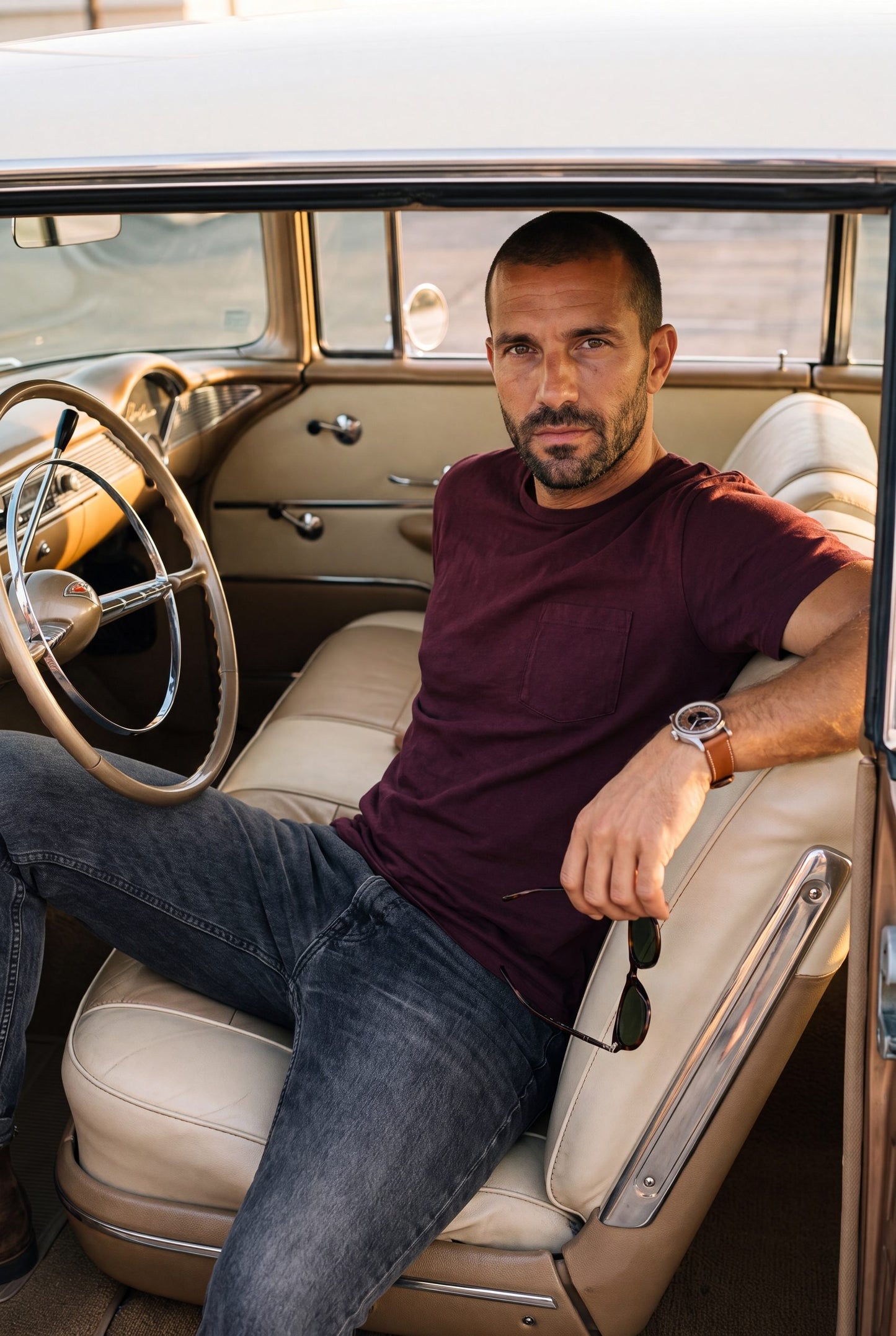 Hombre sentado dentro de un carro clasico vintage con la camiseta de cuello redondo vinotinto con bolsillo de Luck and Load y jeans gris oscuro; foto de estilo de vida.