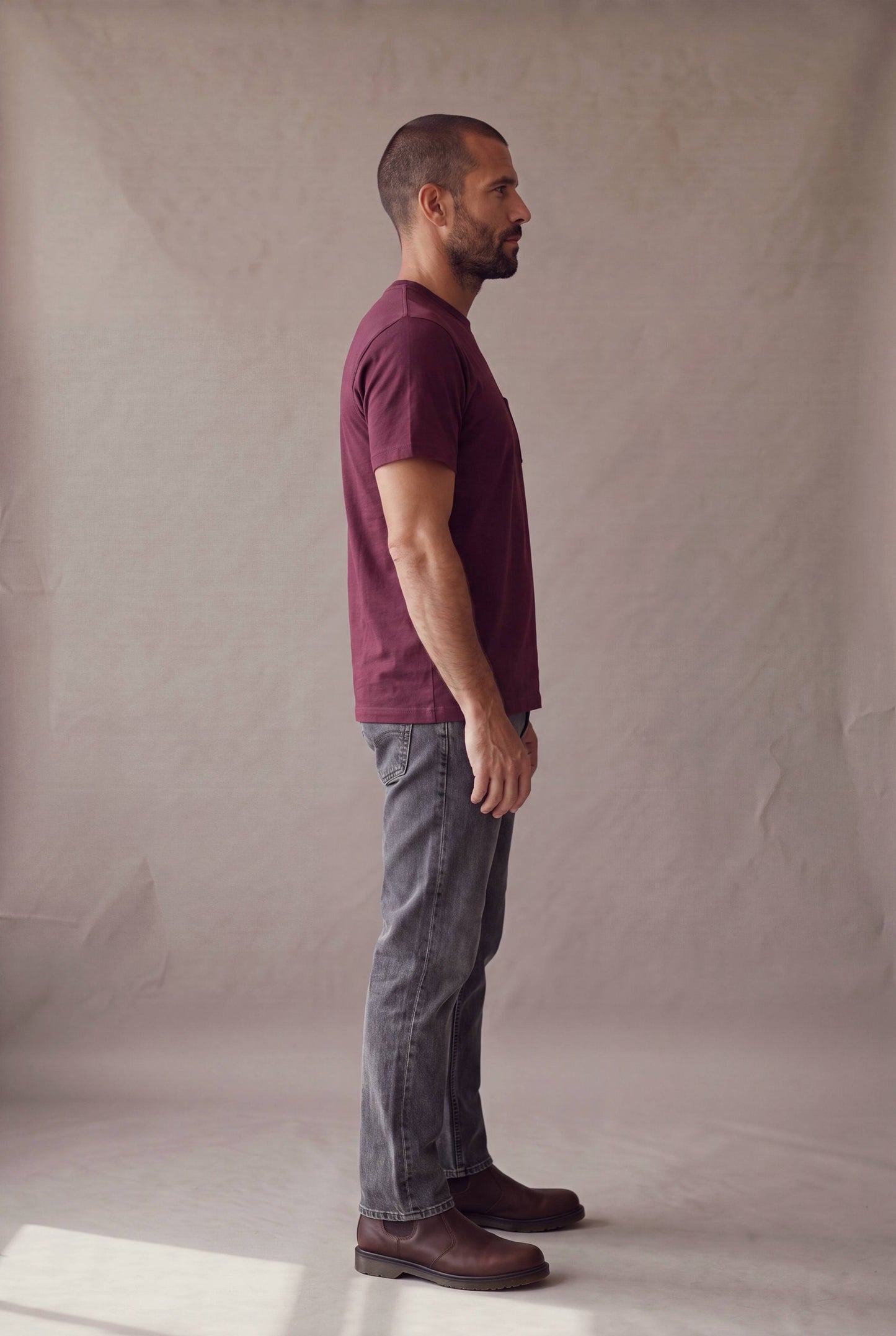 Hombre en perfil derecho con la camiseta de cuello redondo vinotinto con bolsillo de Luck and Load y jeans gris oscuro; foto de catalogo en estudio.