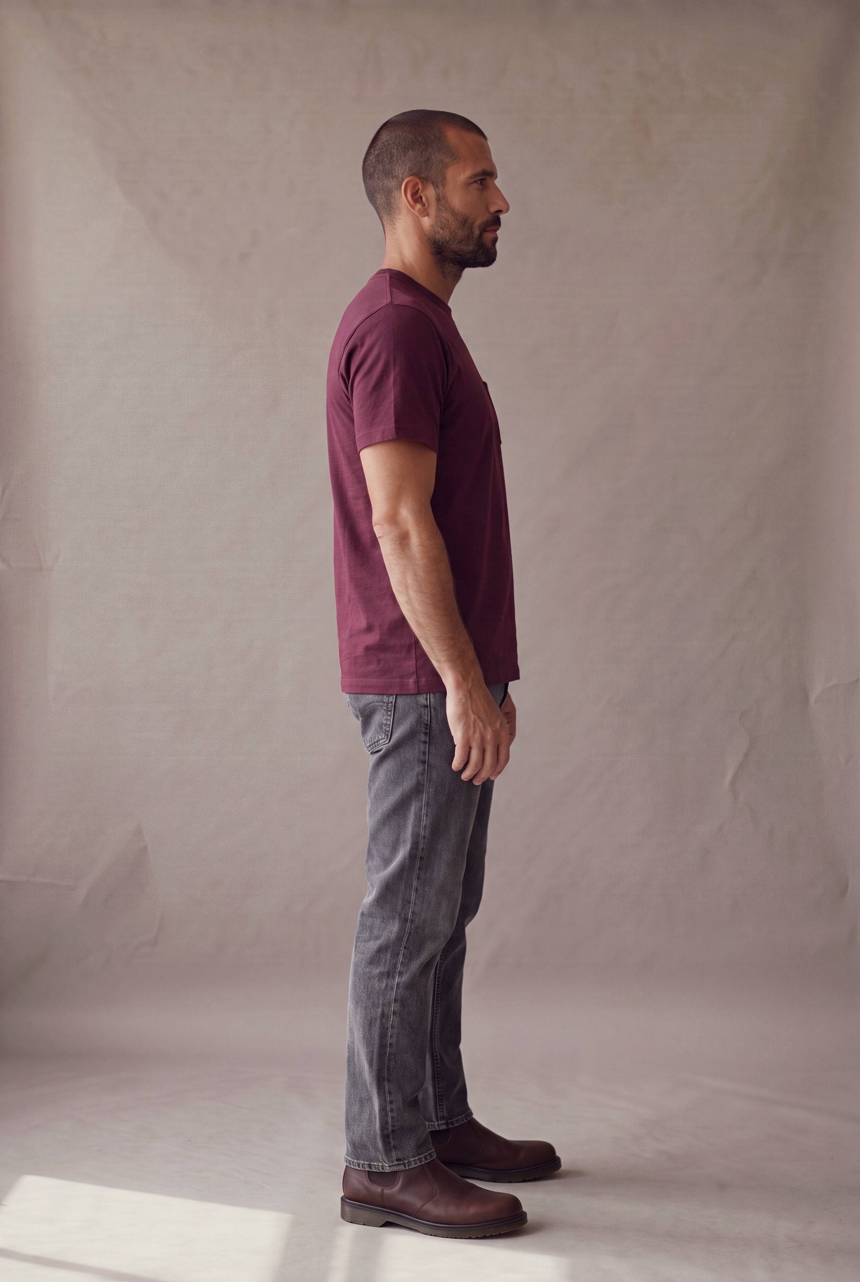 Hombre en perfil derecho con la camiseta de cuello redondo vinotinto con bolsillo de Luck and Load y jeans gris oscuro; foto de catalogo en estudio.