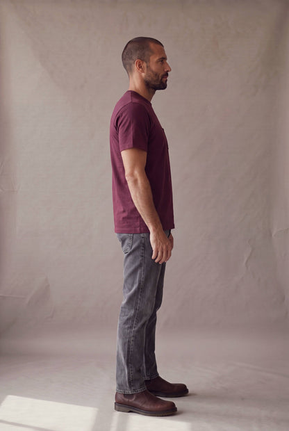 Hombre en perfil derecho con la camiseta de cuello redondo vinotinto con bolsillo de Luck and Load y jeans gris oscuro; foto de catalogo en estudio.