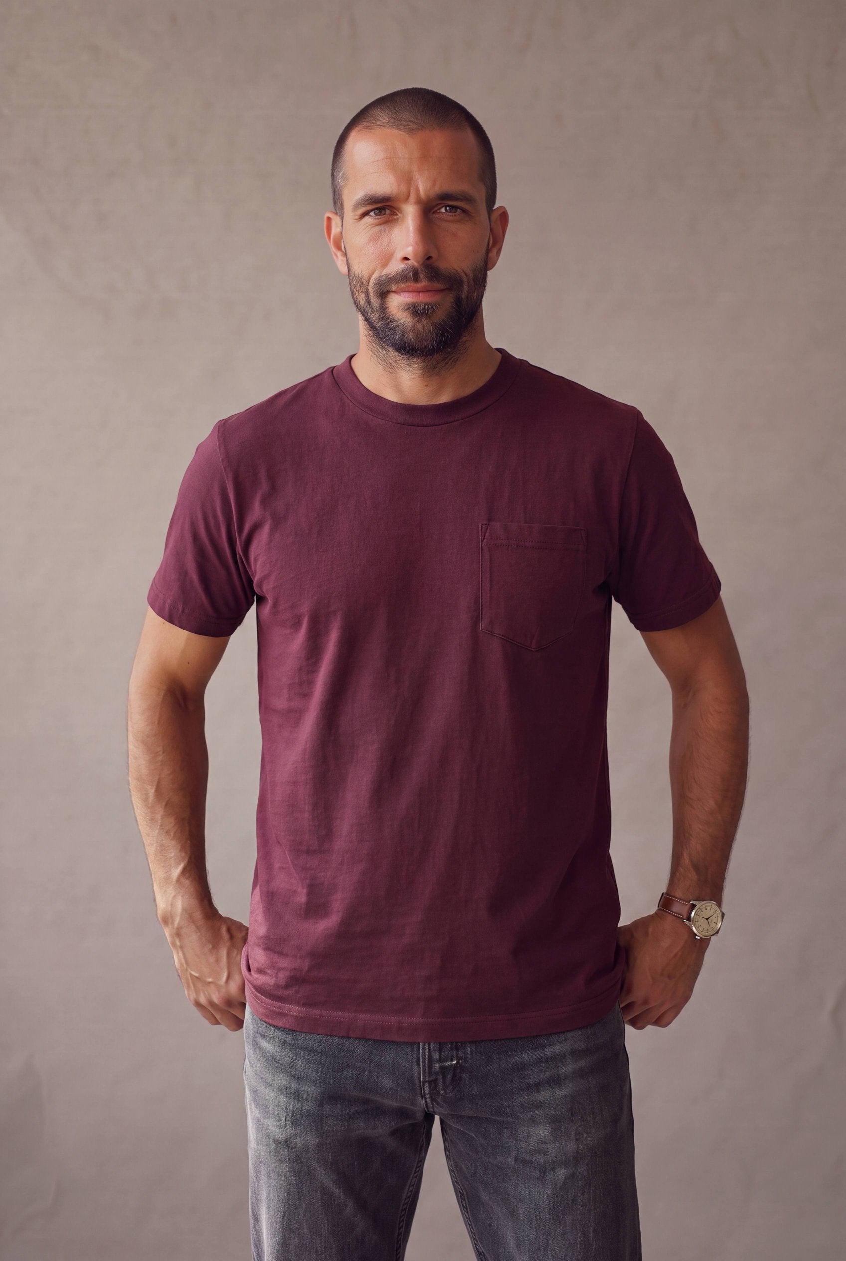 Plano medio de hombre con la camiseta de cuello redondo vinotinto con bolsillo de Luck and Load y jeans gris oscuro; foto de catalogo en estudio.