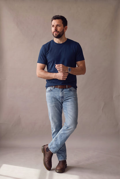 Hombre de pie con camiseta azul marino de cuello redondo sin bolsillo de Luck & Load, con jean claro y botines de cuero café.