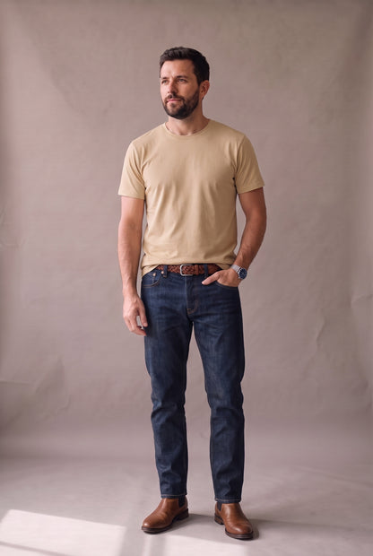 Hombre de pie con camiseta beige de cuello redondo sin bolsillo de Luck & Load, jean índigo oscuro, correa trenzada y botas café, en estudio cálido