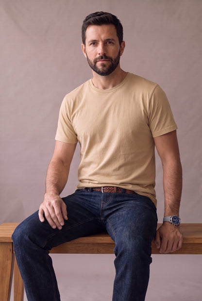Camiseta beige de cuello redondo de Luck & Load, hombre sentado en banca de madera con jean índigo y reloj de acero, medio cuerpo