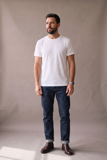 Hombre de pie con camiseta blanca de cuello redondo sin bolsillo de Luck & Load, con jean índigo y botas de cuero café.