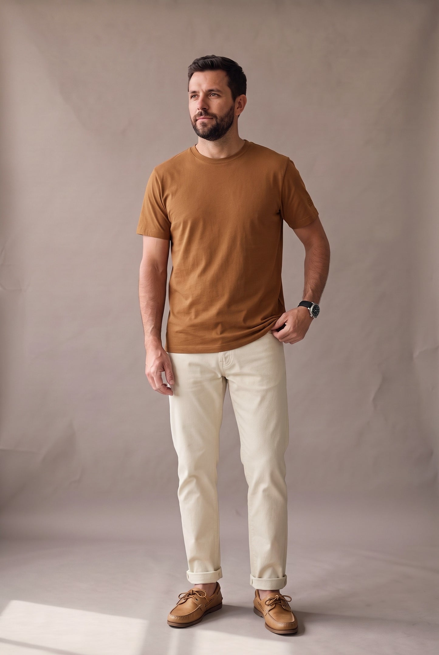 Hombre de pie con camiseta café de cuello redondo sin bolsillo de Luck & Load, pantalón crema y náuticos, en estudio cálido