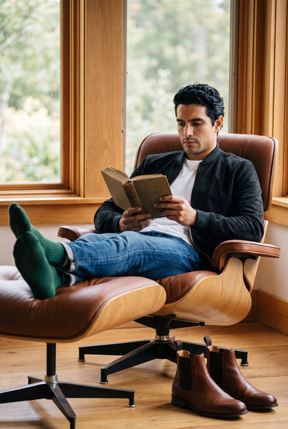 Hombre leyendo en casa en una silla de diseño con la chaqueta bomber negra de gamuza de Luck & Load.