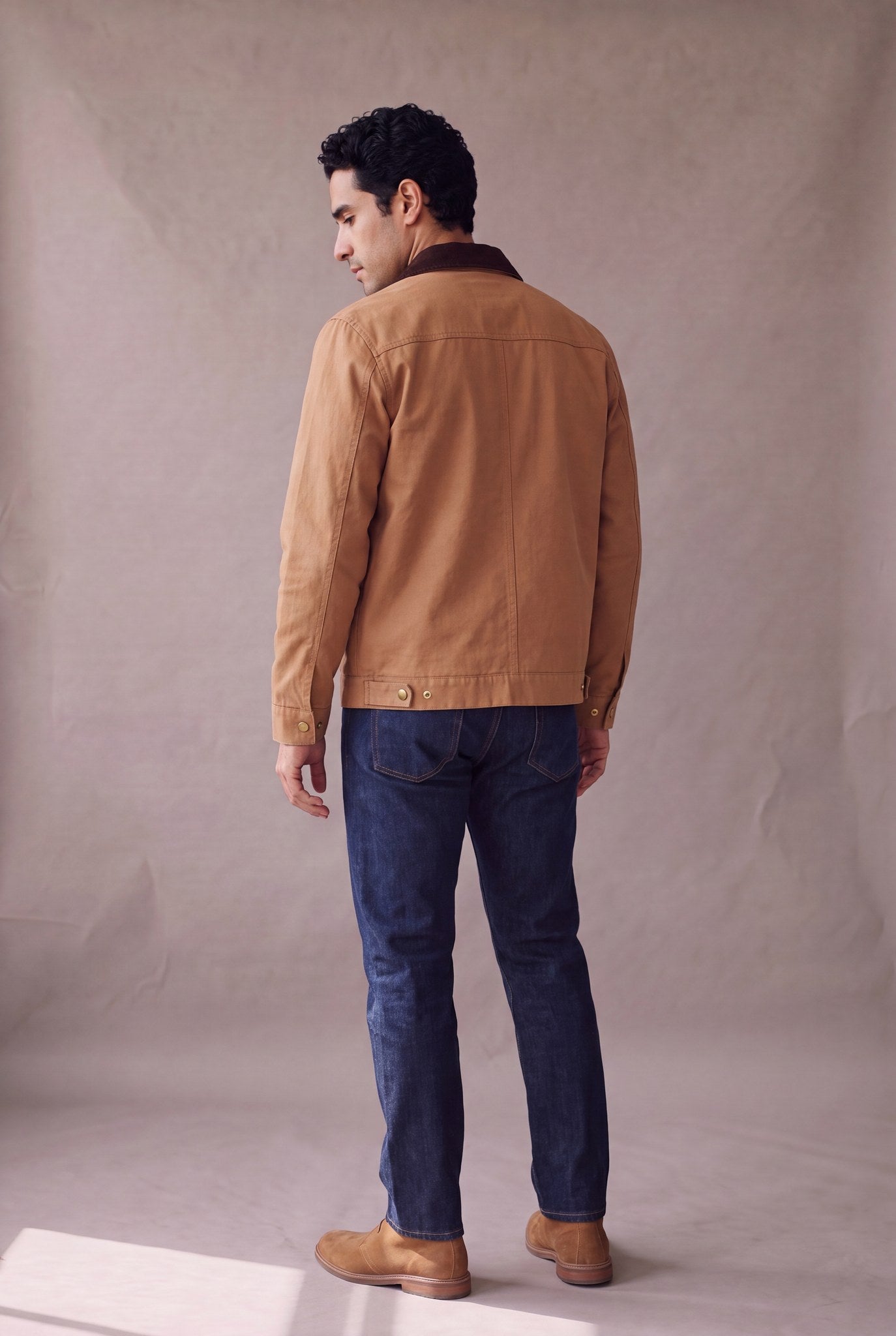 Chaqueta Detroit color camel de Luck and Load en vista de espalda; canesu trasero y broches de laton en el bajo, sobre jean indigo y botas chukka de gamuza. Colombia