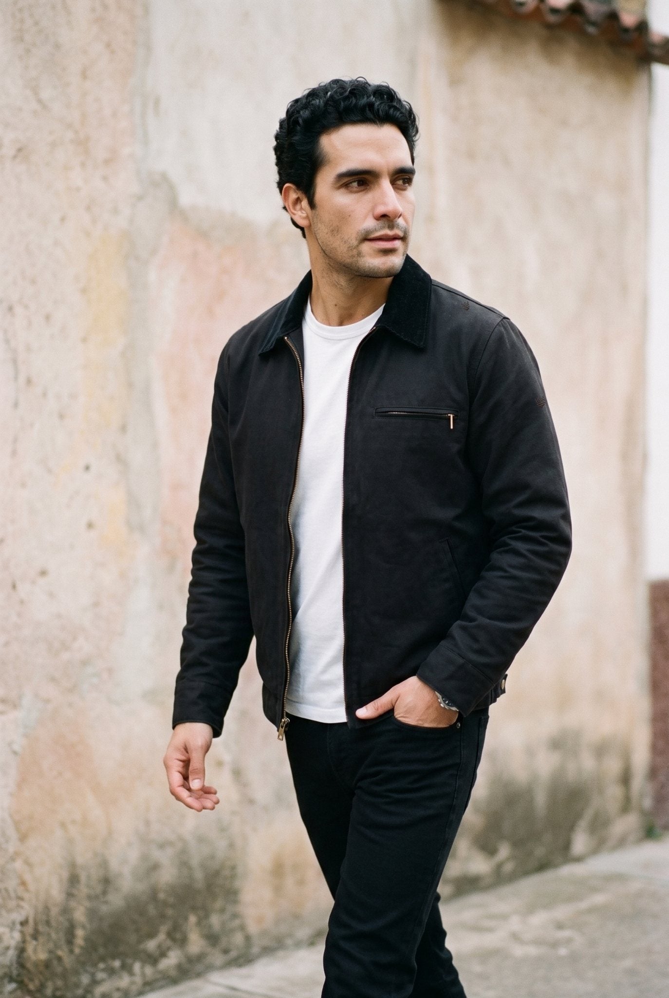 Chaqueta Detroit negra de cuello de pana de Luck and Load; hombre caminando junto a un muro de calle de dia, plano americano, camiseta blanca y jean negro recto. Colombia