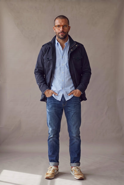 Hombre con chaqueta militar azul marino Luck and Load abierta sobre camisa oxford celeste, jean y tenis, de frente