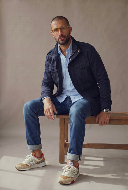 Hombre sentado con chaqueta militar azul marino Luck and Load, camisa oxford celeste y jean, en banca de estudio