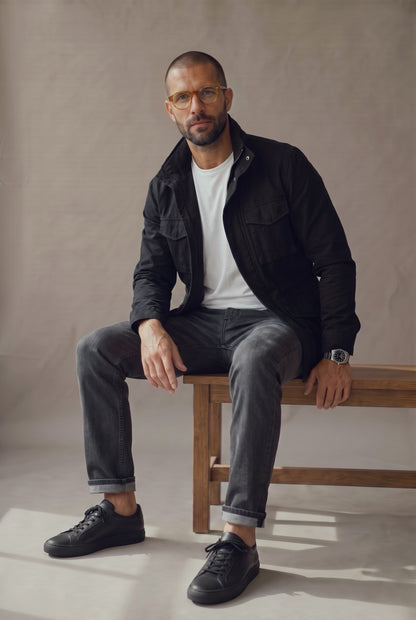 Hombre sentado con chaqueta militar negra Luck and Load, camiseta blanca y jean gris, en banca de estudio