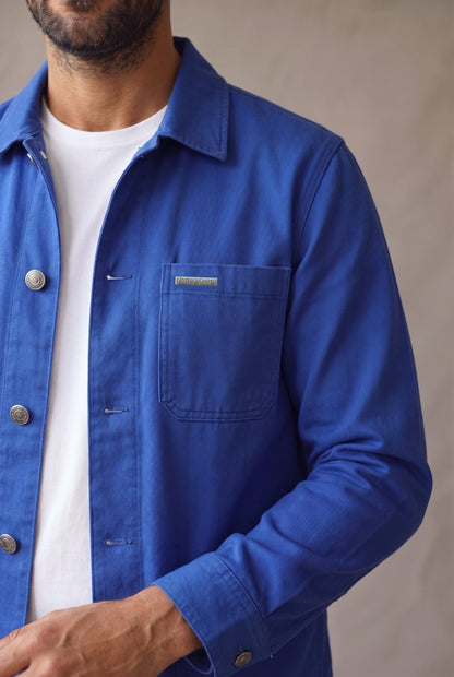 Detalle de la chaqueta obrera azul rey de Luck & Load: bolsillo de pecho con placa metálica grabada, botones de peltre y ojales horizontales sobre sarga de algodón.