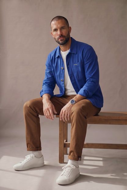Chaqueta obrera azul rey para hombre de Luck & Load, sentado en una banca de madera con las mangas arremangadas y pantalón café.