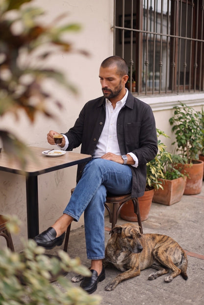 Hombre con chaqueta obrera negra de Luck & Load sentado en el patio de un café con su perro, foto de estilo de vida en exteriores.