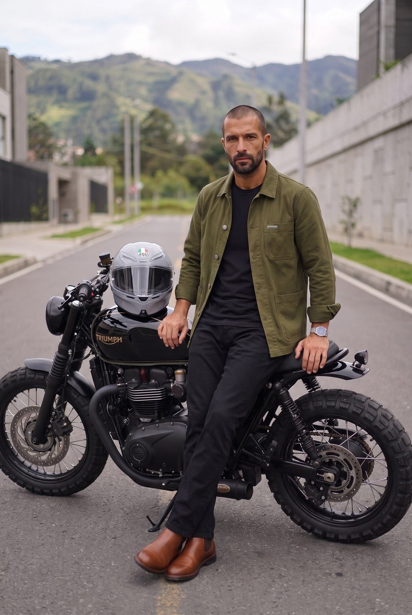 Hombre con chaqueta obrera verde militar de Luck & Load recostado contra su moto en una calle de ladera con los cerros verdes al fondo.