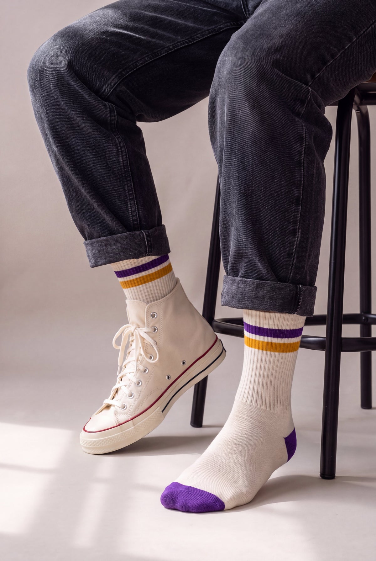 Medias largas crema con franja morada y dorada, en hombre sentado con jean negro y Converse crema, caña completa a la vista