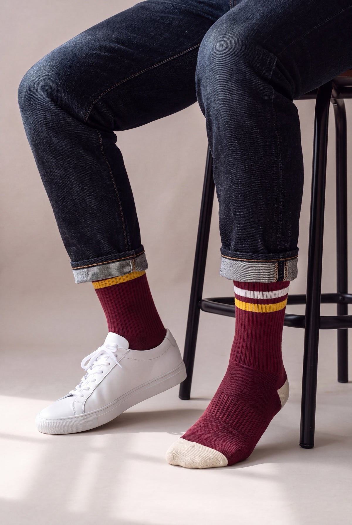 Medias largas vinotinto con franja dorada, en hombre sentado con jean oscuro y tenis blancos Common Projects